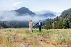 a man and woman holding hands while walking through a field with mountains in the background