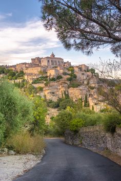 an old village on top of a hill with trees in the foreground and a paved road leading up to it