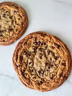 two chocolate chip cookies sitting on top of a white marble counter next to each other