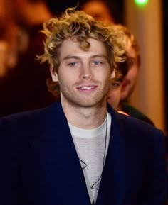 a man with curly hair is smiling at the camera while wearing a blue suit and white t - shirt