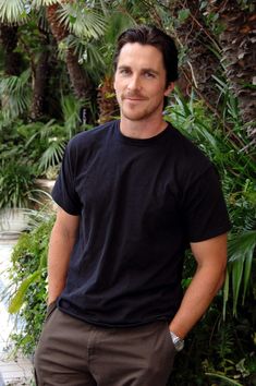 a man in black shirt and brown pants standing next to palm trees with his hands on his hips