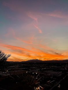 an orange and pink sunset with clouds in the sky above some buildings, trees and mountains