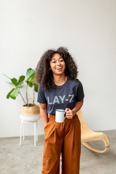 a woman holding a coffee cup and smiling at the camera while standing in front of a plant