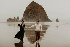 an engaged couple holding hands and walking on the beach in front of some rock formations