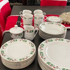 a table topped with lots of white plates and cups covered in holly designs next to a red chair