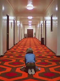 a young boy sitting on the floor in front of an orange and red carpeted hallway