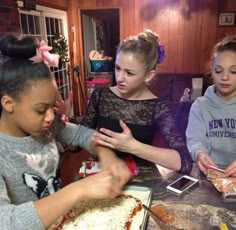 two girls are helping another girl put toppings on a pizza crust in the kitchen