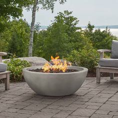 an outdoor fire pit sitting on top of a brick patio next to chairs and trees