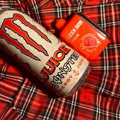 an orange and white drink sitting on top of a red plaid bed spread next to a can of bud light