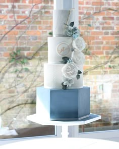 a three tiered blue and white wedding cake with flowers on the top, sitting in front of a window