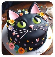 a black cat cake with flowers and butterflies on it's face is sitting on a plate