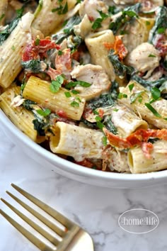 a white bowl filled with pasta covered in chicken and spinach sauce next to a gold fork