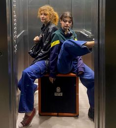 two people sitting on top of a piece of luggage in an elevator stall with the doors open