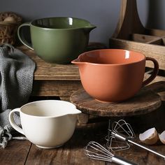 various kitchen utensils are sitting on a wooden table next to bowls and whisks