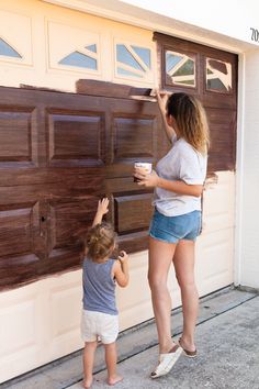 a woman and child are painting the side of a garage door with brown paint on it