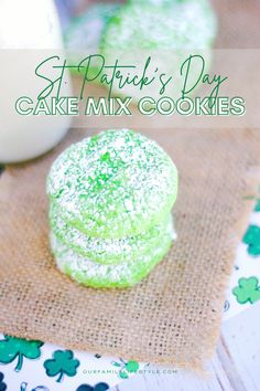 stack of three light green cake mix cookies with white powered sugar and green crystals stacked on a burlap piece of fabric overlaying a white placemat with green shamrocks. A gallon milk and more cookies are blurred in the background.