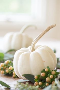 two white pumpkins sitting on top of a table next to green leaves and berries