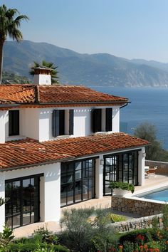 a white house with red tile roofing next to the ocean and palm trees in front of it