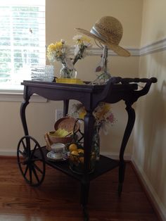 a small cart with flowers and hats on top in front of a window next to a wooden floor