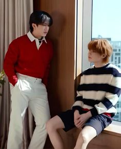 two young men sitting next to each other in front of a window, one wearing a red sweater and the other white pants