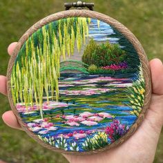 a person holding up a small embroidery art piece with water lilies and willow trees in the background