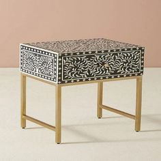 a small black and white table with gold trimmings on the top, in front of a pink wall