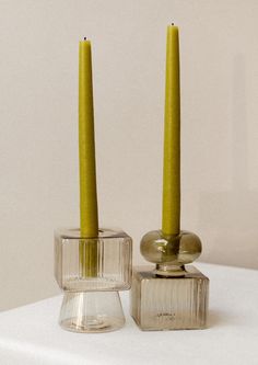 two candles are placed in glass holders on a white tablecloth, one is yellow and the other is clear