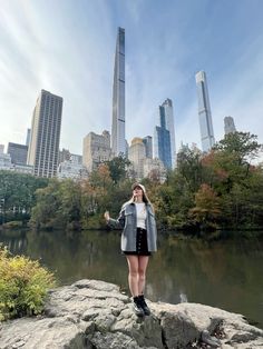 a woman standing on top of a rock next to a lake in front of tall buildings