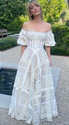 a woman standing in front of a pond wearing a white dress with an off the shoulder design