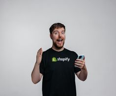 MrBeast holds a cell phone, looking surprised, in a black t-shirt featuring the Shopify logo