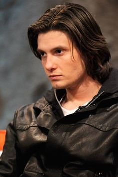 a man with long hair wearing a black leather jacket and white t - shirt sitting down