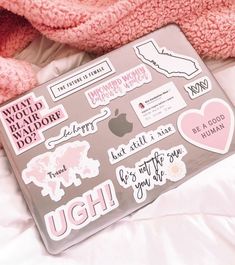 a laptop computer covered in stickers on top of a white bed next to a pink teddy bear