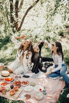 two women sitting on a blanket in the woods having a picnic with food and drinks