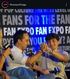 two people sitting on a stage with microphones in their hands and one person standing up