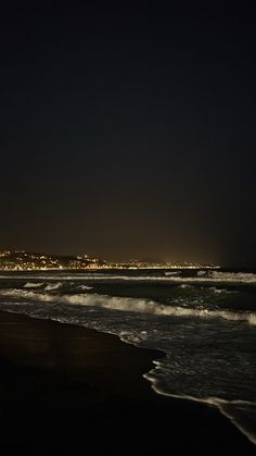 the beach is dark with waves coming in to shore and buildings lit up at night