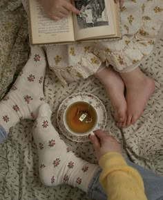 two people sitting on a bed with an open book and a cup of tea in front of them