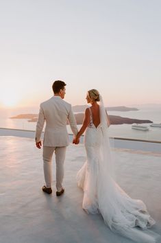 a bride and groom holding hands while standing in front of the ocean at sunset on their wedding day