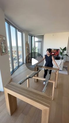 a woman standing on top of a wooden table in a living room next to large windows