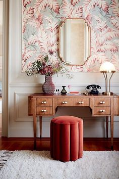 a desk with a mirror, stool and flowers on it in front of a wall