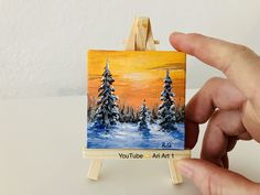 someone is holding up a miniature painting on a small easel with trees in the background