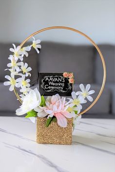 a white and pink flower arrangement in a gold ring with a black sign that says awn's community