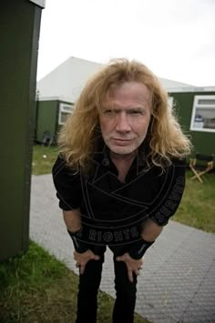 a man with long blonde hair standing in front of a green building and looking at the camera