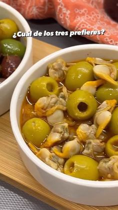 two white bowls filled with green olives on top of a wooden cutting board