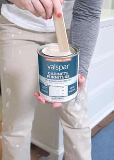 a woman holding a can of paint and a wooden stick in her hand while standing on the floor