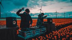 two men in a field with windmills behind them and one man playing dj's