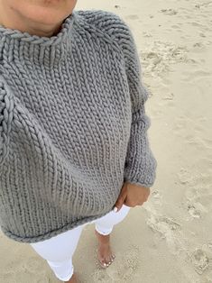 a woman standing on top of a sandy beach wearing a sweater and white pants with her hands in her pockets