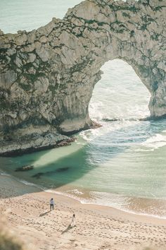 two people walking on the beach in front of an arch shaped rock formation near the ocean