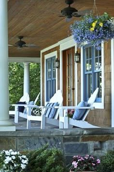 a porch with blue chairs and flowers hanging from it's ceiling over the front door