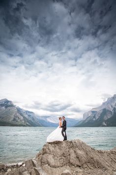 a bride and groom standing on top of a rock in front of the water with mountains behind them