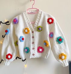 a white cardigan with colorful flowers on it hanging from a clothes hook, next to a string of lights